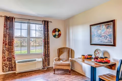 A cozy corner of a room with a large window covered by floral curtains, a beige upholstered armchair, a wooden table set with bowls and plates, a chair, and a framed painting on the wall.