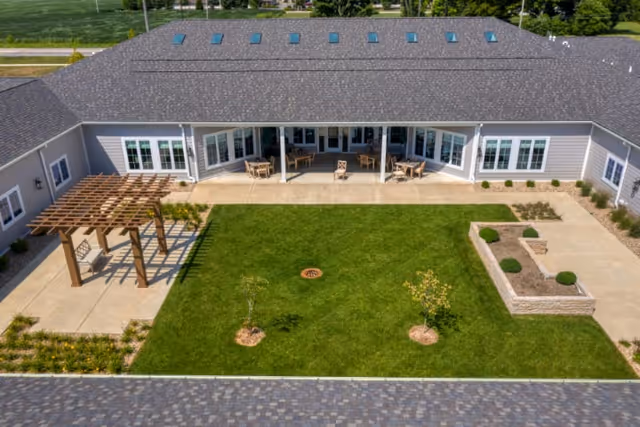 Aerial view of a senior living facility courtyard with a well-maintained green lawn, small trees, a wooden pergola with seating, and a covered patio area with tables and chairs attached to a single-story building with multiple windows and skylights on the roof.