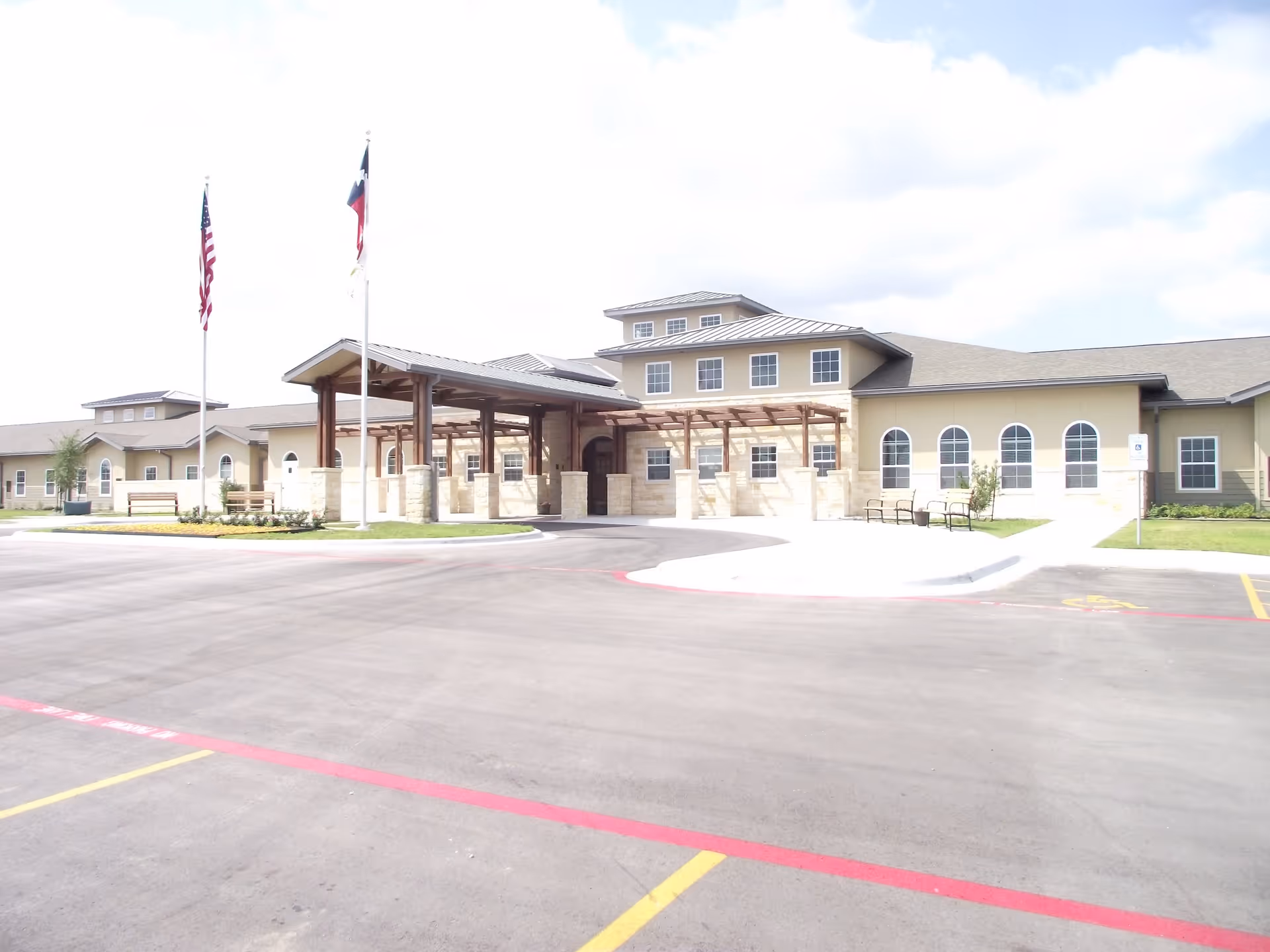 Exterior view of Stoney Brook of Copperas Cove senior living facility showing a large building with beige walls, multiple windows, and a covered entrance supported by wooden beams. Two flagpoles with American and Texas flags are visible near the entrance. The foreground shows a paved parking area with marked spaces and a red curb.