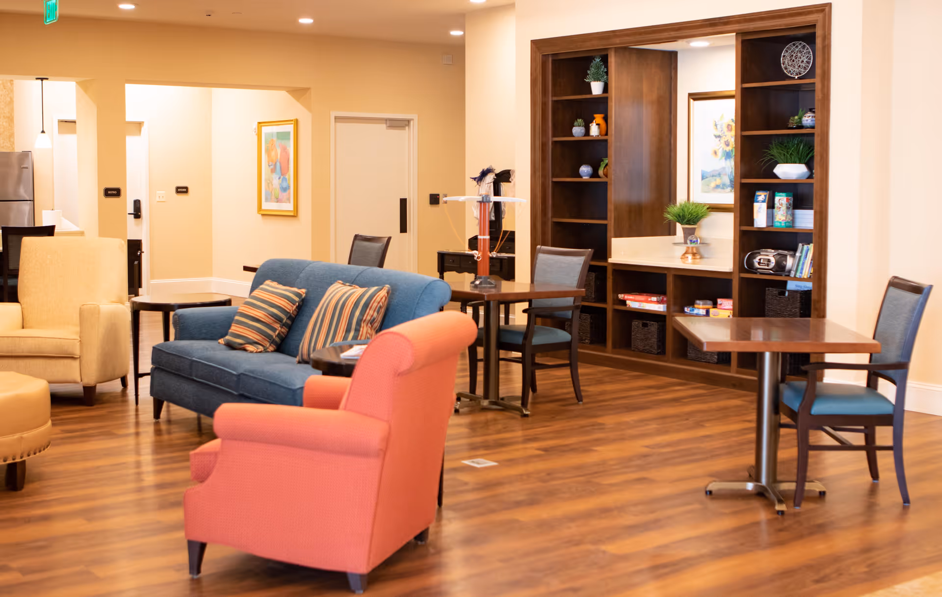 A cozy common area with wooden flooring featuring a blue sofa with striped cushions, a coral armchair, a beige armchair, and small wooden tables with chairs. There is a built-in wooden shelving unit with decorative items and a framed picture on the wall. The space is warmly lit with recessed ceiling lights.