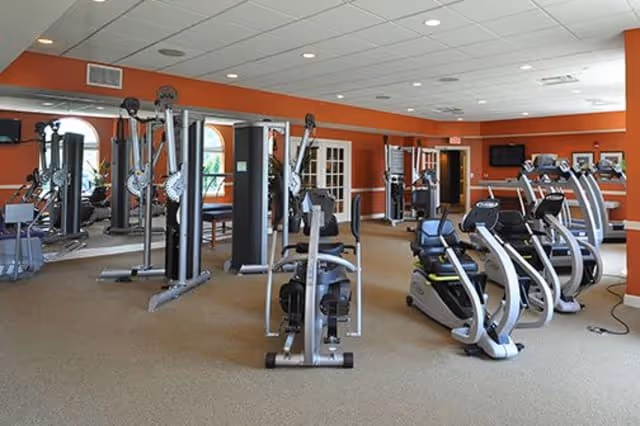 Interior view of a fitness room in a senior living facility with various exercise machines including stationary bikes, weight machines, and cardio equipment. The room has orange walls, a beige carpet, and a ceiling with recessed lighting.
