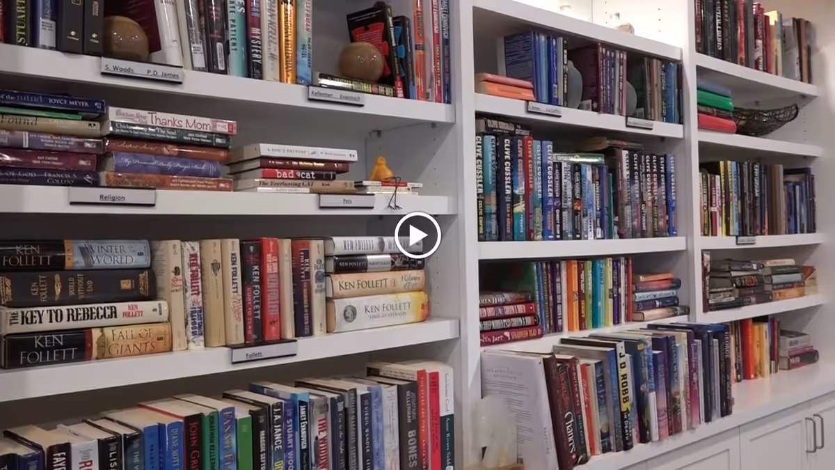 Bookshelves filled with a variety of books organized by categories such as Religion, Pets, and Follett, in a well-lit room with white shelves and cabinets below.