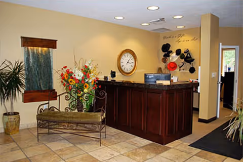 Reception area with a dark wooden front desk, a large round wall clock, and a decorative wall-mounted hat rack with various hats. There is a bench with a floral arrangement in front of it, a potted plant to the left, and a water feature on the wall. The floor is tiled and the walls are painted beige.