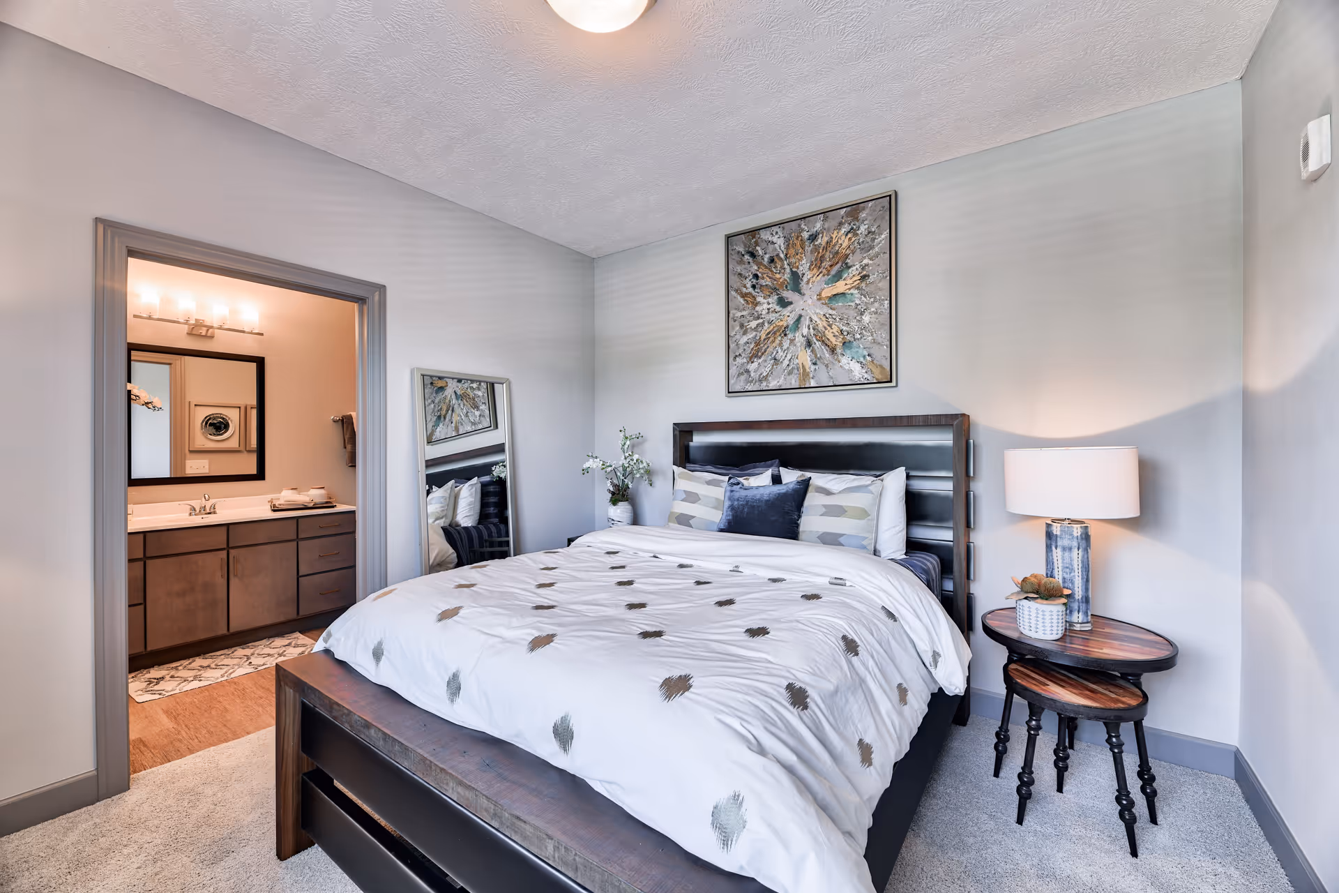 A modern bedroom with a large bed featuring a white comforter with a dotted pattern and multiple pillows. There is a bedside table with a lamp and a small plant. A large abstract painting hangs above the bed. A full-length mirror leans against the wall near the entrance to an adjoining bathroom, which has a vanity with a sink, a mirror, and lighting fixtures.