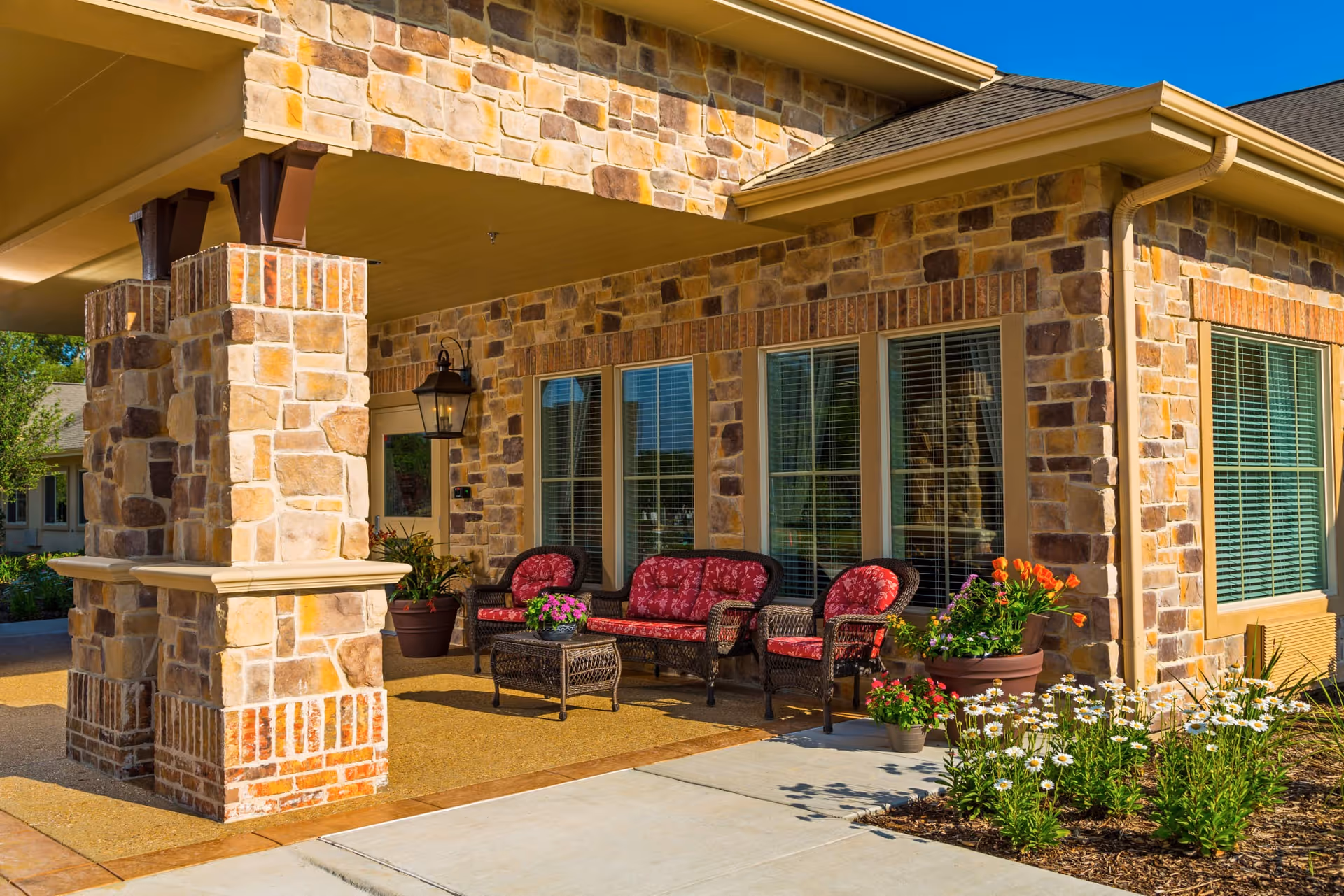 Outdoor seating area at Riverside Oxford Memory Care featuring stone pillars and walls, a covered patio with wicker furniture including a sofa and two chairs with red cushions, a small table with a flower pot, and surrounding flower beds with various plants and flowers.