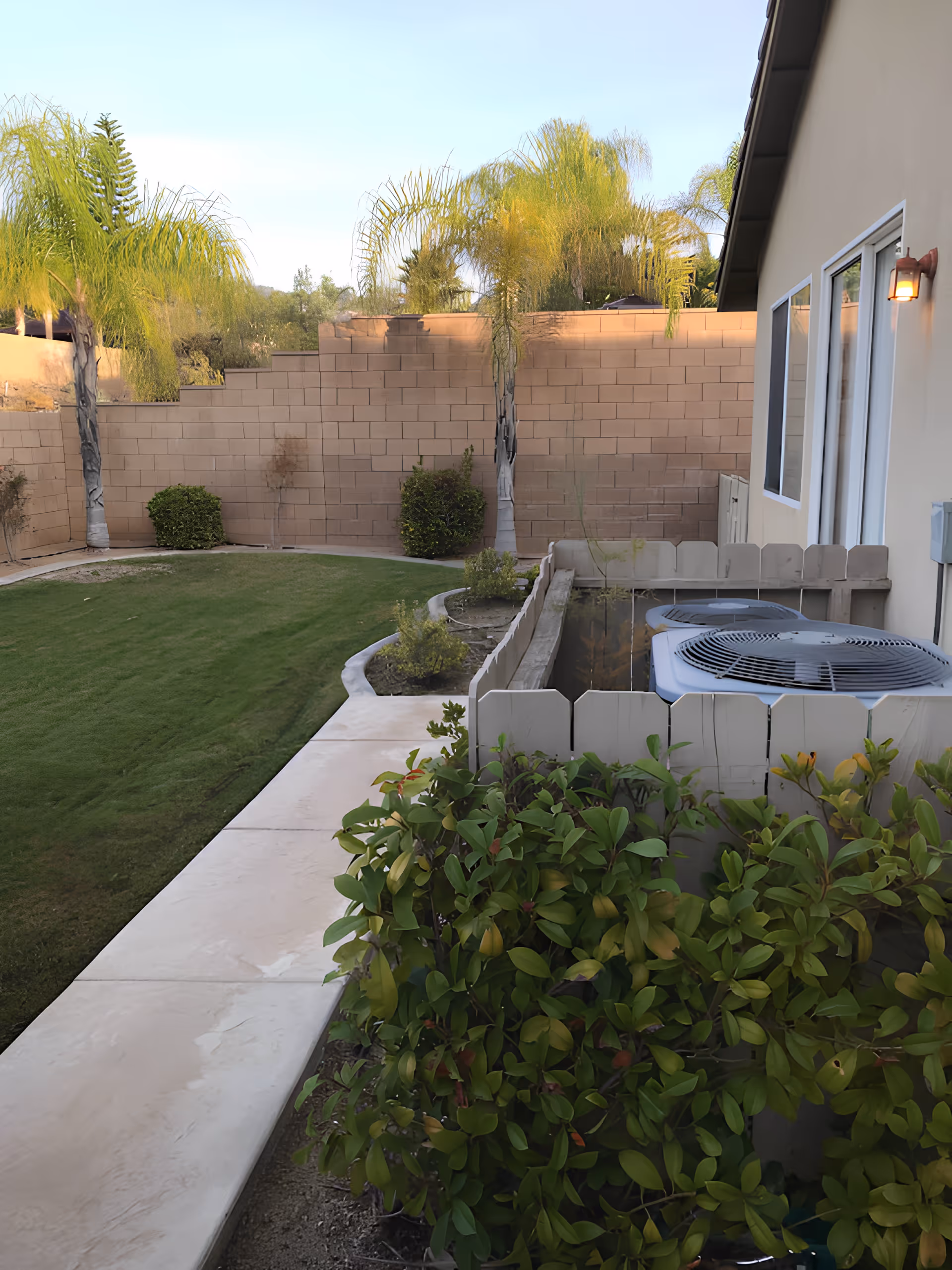 Outdoor view of a backyard area with a concrete walkway, green grass lawn, palm trees, bushes, and a beige brick wall. On the right side, there is the exterior wall of a building with windows and an outdoor light fixture, along with two air conditioning units enclosed by a white wooden fence.
