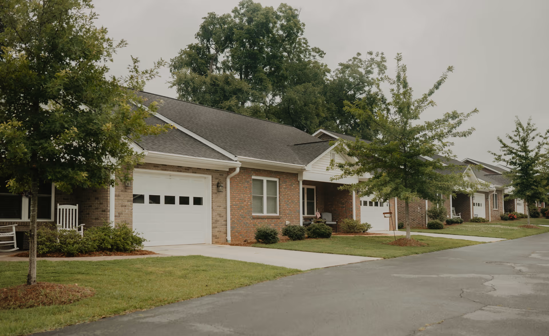 Row of single-story brick residential units with garages, small front porches, and green lawns with young trees along a paved road under an overcast sky.