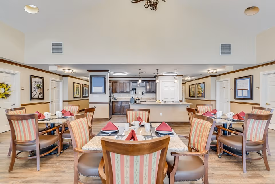A dining area in a senior living facility with several tables set for meals. Each table has four chairs with striped upholstery and place settings including red folded napkins, white mugs, and plates. In the background, there is a kitchen area with dark wooden cabinets, a countertop, and hanging pendant lights. The walls are decorated with framed artwork and wreaths, and the floor is wood.