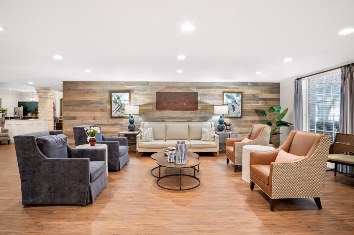 A cozy living room area in Appletree Court featuring a beige sofa with two patterned pillows, two blue armchairs with a small round table holding a vase of flowers, and two brown armchairs with a small white side table between them. The room has wooden flooring, a rustic wooden accent wall with two framed botanical prints and a sign that reads 'Appletree Court'. Two blue lamps are placed on side tables flanking the sofa. A large window with curtains allows natural light into the space.