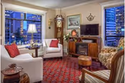 A cozy living room with a white sofa, two armchairs, a wooden grandfather clock, a wooden cabinet with a TV on top, and a red patterned carpet. Large windows show a cityscape at dusk with tall buildings and lights.