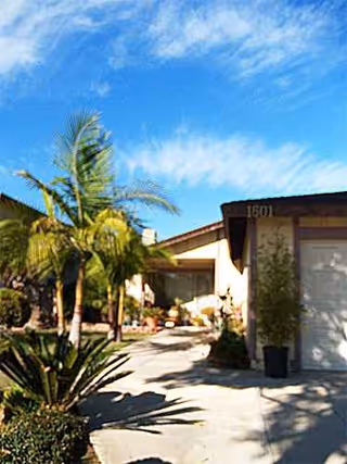 Front exterior of a senior living home with palm trees and a driveway.