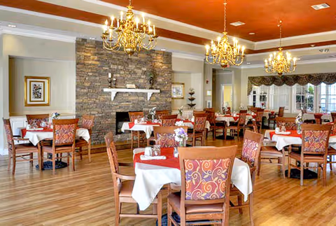A spacious dining room with multiple tables covered in white and red tablecloths, each set with dining utensils and small flower arrangements. The room features wooden chairs with patterned upholstery, a stone fireplace in the background, and elegant chandeliers hanging from the ceiling. Large windows with decorative curtains allow natural light to fill the space.