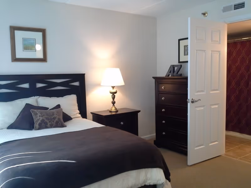 A bedroom with a neatly made bed featuring white and dark brown pillows and a dark brown blanket. Next to the bed is a nightstand with a lit table lamp. Across from the bed is a tall dark wooden dresser with decorative items on top. The door to the room is open, revealing a hallway with patterned wallpaper.