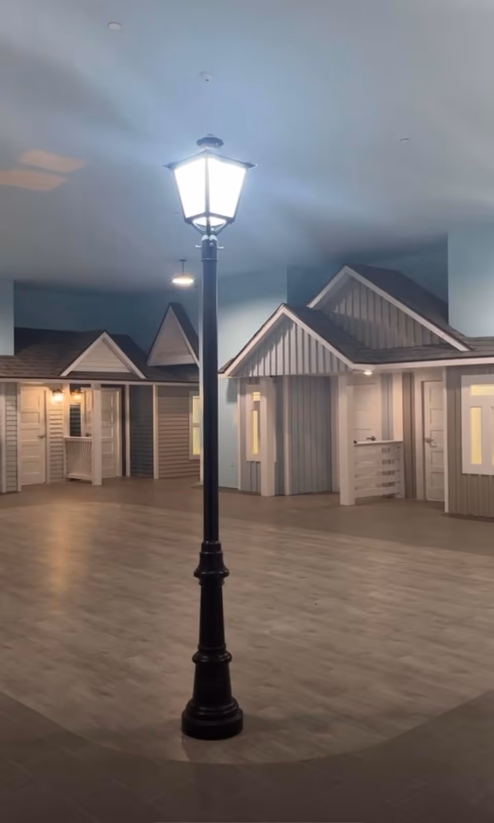 An indoor common area featuring a tall black lamppost surrounded by small cottage-style room facades and wide flooring.