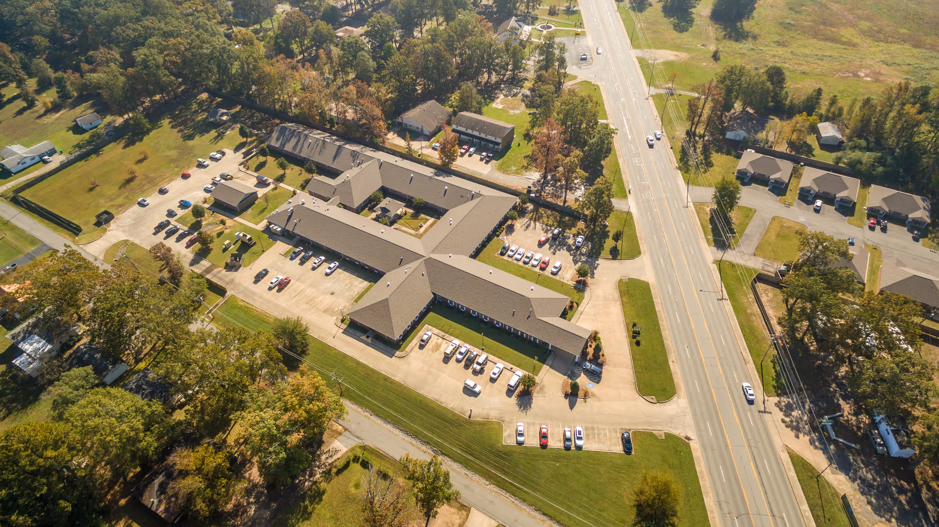 Aerial view of a single-story senior care facility with multiple connected wings, parking lots, lawns, trees, and a main road alongside.
