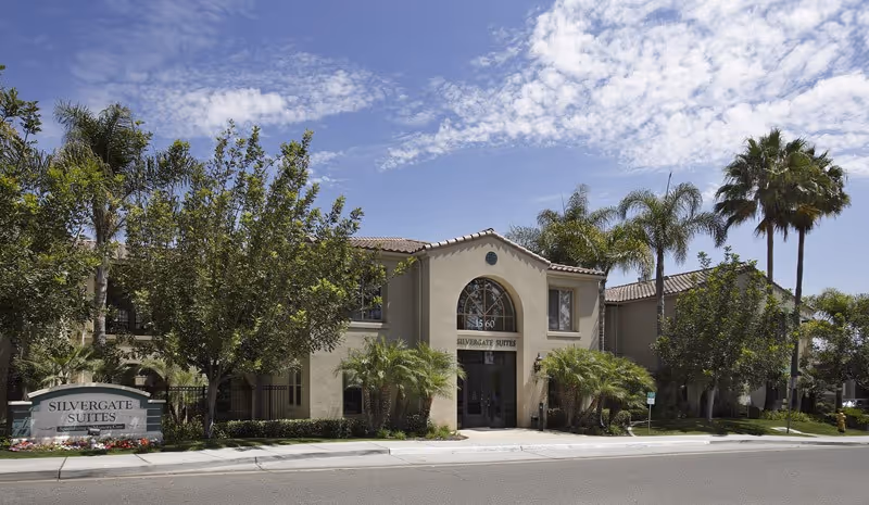 Front exterior of the Silvergate Suites building with an arched entrance, palm trees, and a sign by the sidewalk.