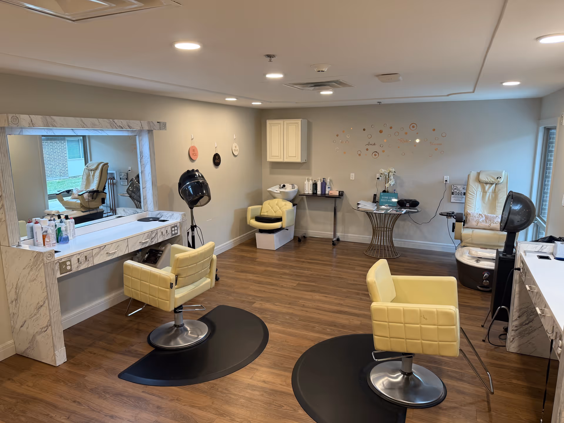 A bright salon room with wooden flooring featuring two yellow salon chairs on black mats facing a large mirror with a marble frame. There is a hair dryer hood, a small yellow chair near a wash basin, and a pedicure chair with a foot bath near a window. Various hair and beauty products are placed on counters and a small table, with wall decorations and ceiling lights illuminating the space.