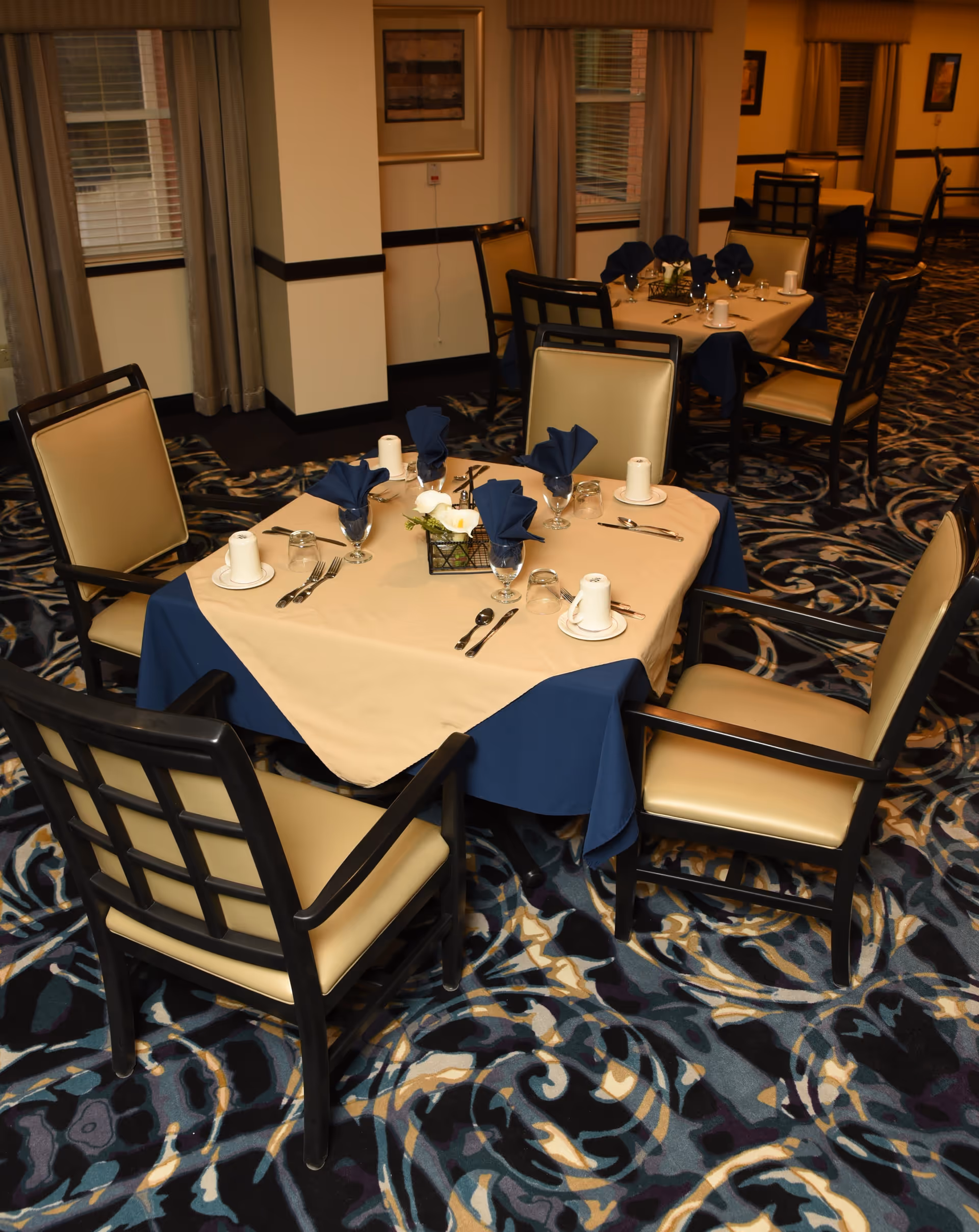 Square dining table set for four with navy napkins, glassware, and place settings in a carpeted communal dining room.