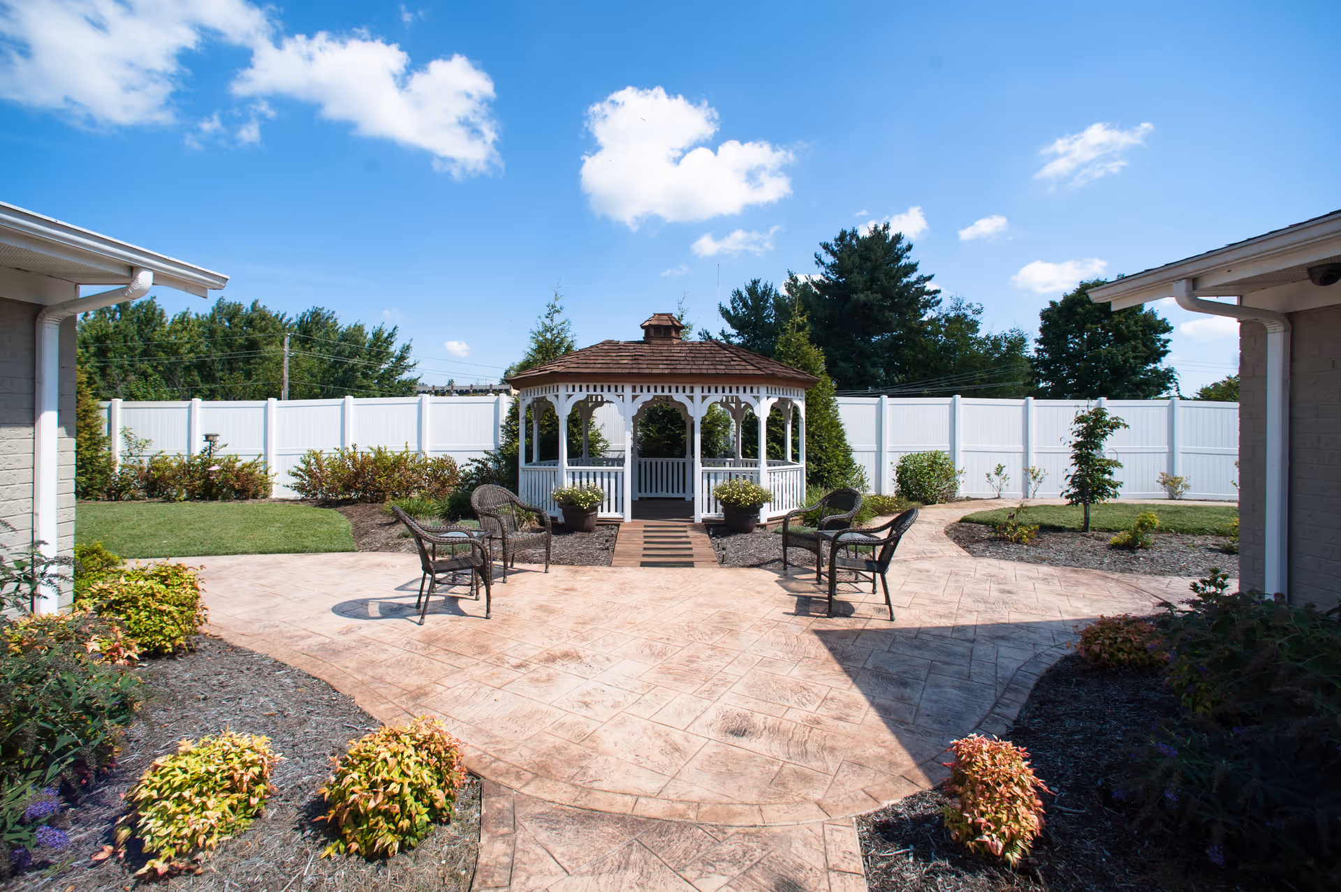 Outdoor courtyard area with a white gazebo in the center, surrounded by a paved walkway and several black wicker chairs. The area is landscaped with small bushes and plants, enclosed by a white fence, under a blue sky with some clouds.