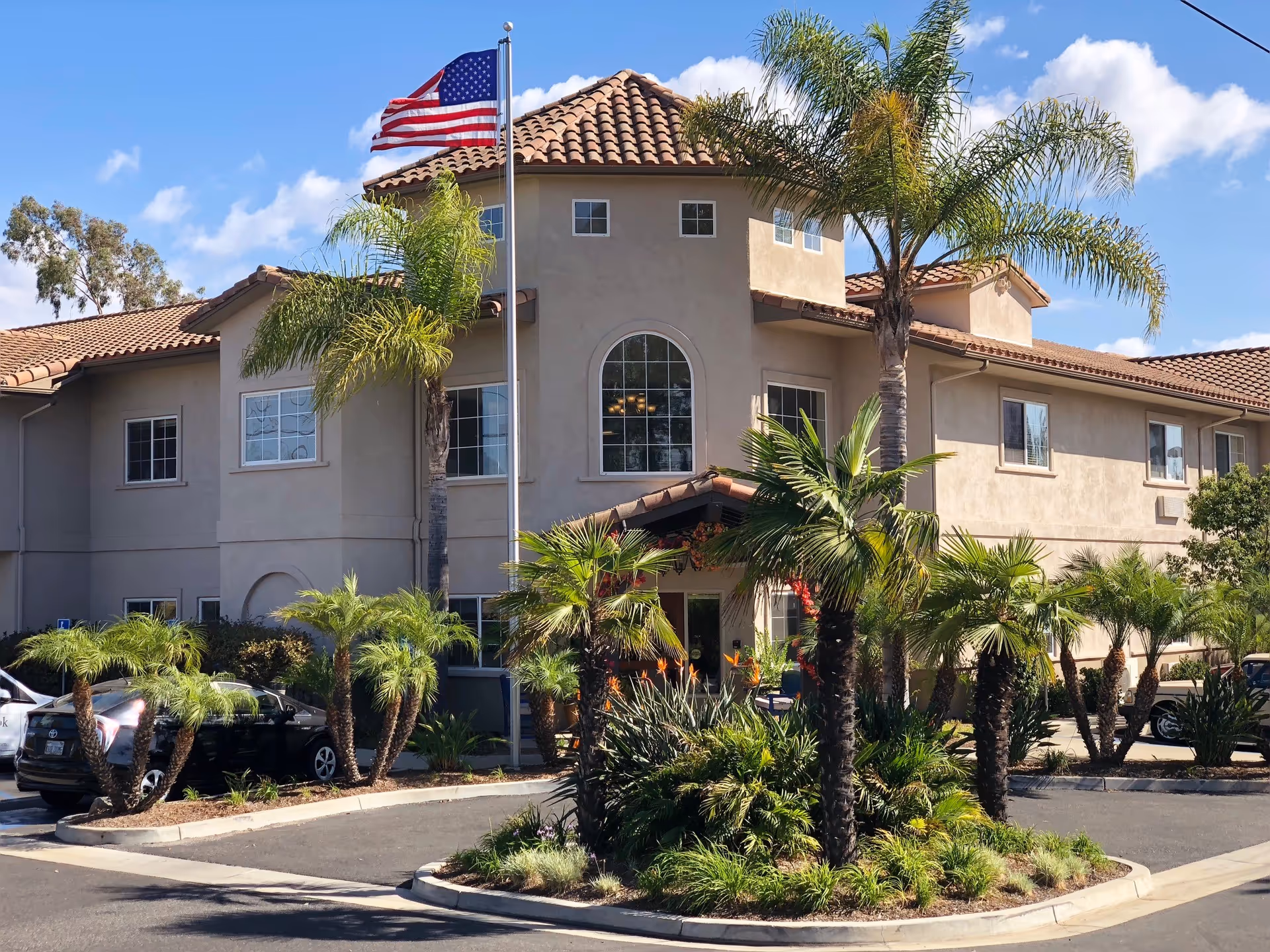 Exterior view of a two-story building with a tiled roof and beige walls, surrounded by palm trees and landscaping. An American flag is flying on a flagpole in front of the building under a clear blue sky.