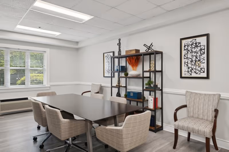 A modern meeting or dining room with a rectangular dark table surrounded by six beige upholstered chairs on wheels. The room has light gray walls with white trim, a large window with blinds, and a black metal shelving unit decorated with various items including vases, books, and plants. Two abstract black and white framed artworks hang on the wall.