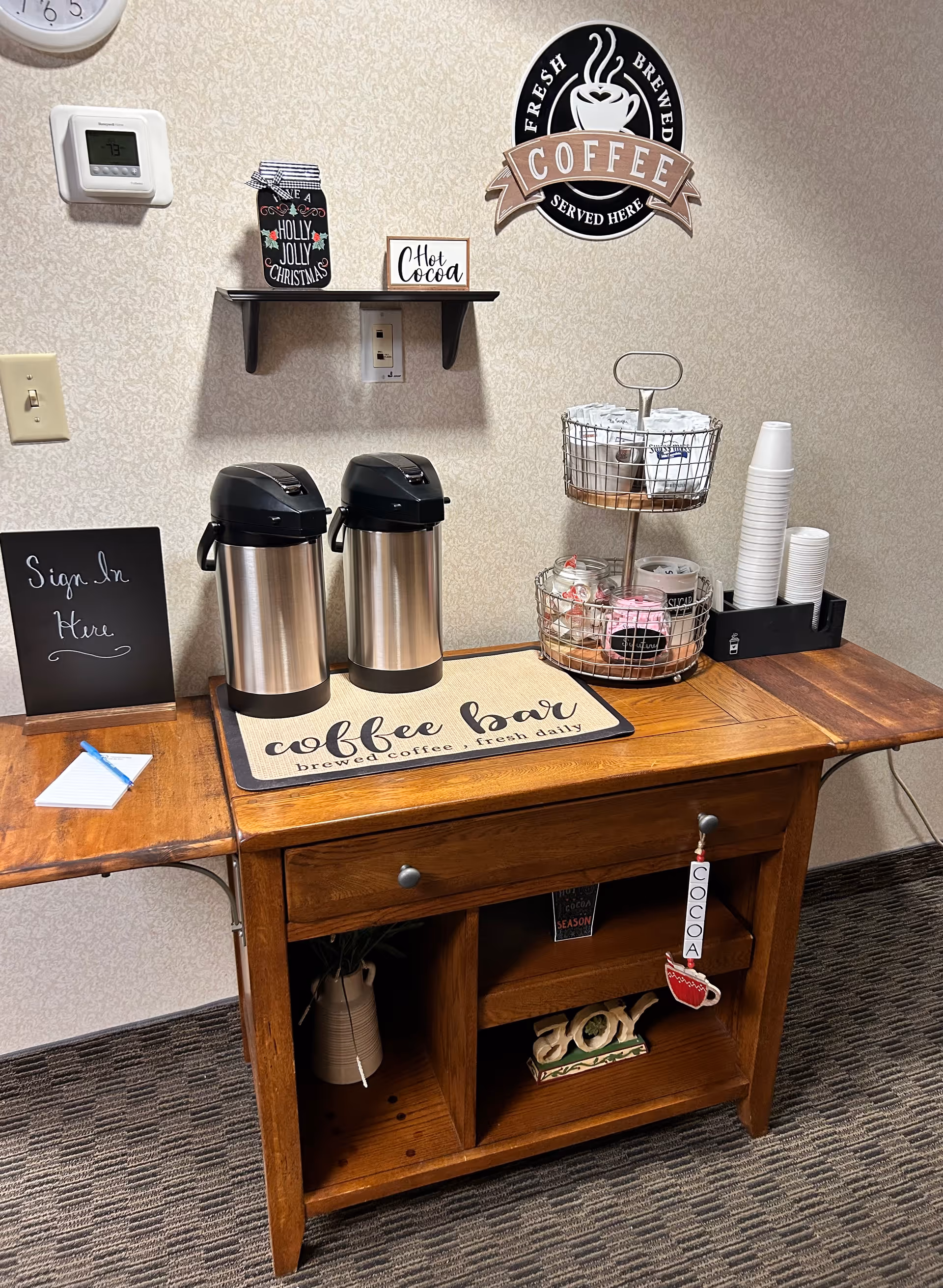 A coffee bar setup on a wooden table with two stainless steel airpots, a two-tiered wire basket holding coffee supplies, a stack of disposable cups, and a small chalkboard sign that says 'Sign In Here' with a notepad and pen. The wall behind has a thermostat, a clock, a small shelf with holiday-themed signs, and a round sign that reads 'Fresh Brewed Coffee Served Here.'