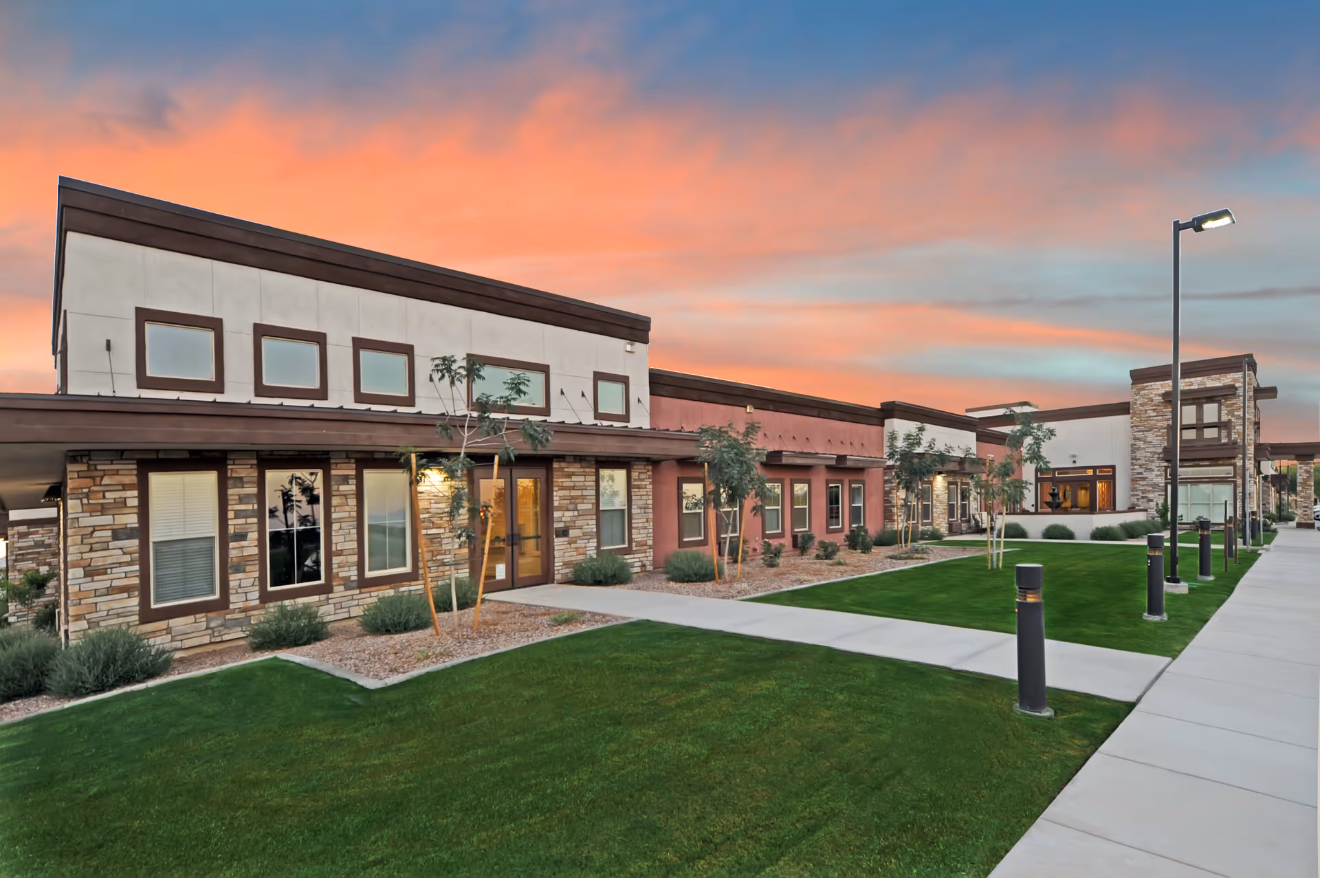 Exterior view of a modern senior living facility building at sunset with a well-maintained lawn, small trees, and a paved walkway with lamp posts.