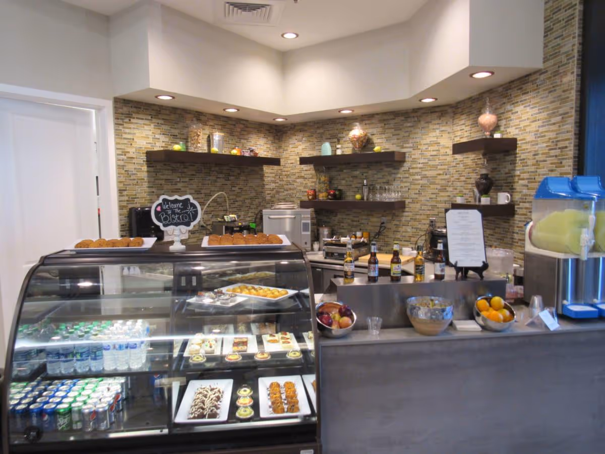 Small café/bistro counter with a glass refrigerated display of pastries and drinks, shelves with decorative items, and a beverage station.
