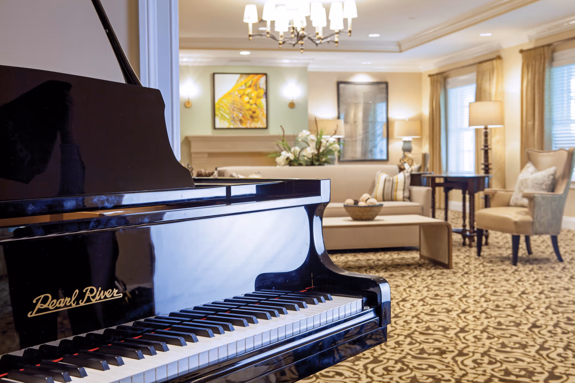A polished black Pearl River grand piano in the foreground with a cozy living room in the background featuring a beige sofa, armchairs, a coffee table with decorative items, lamps, framed artwork on the walls, and patterned carpet.