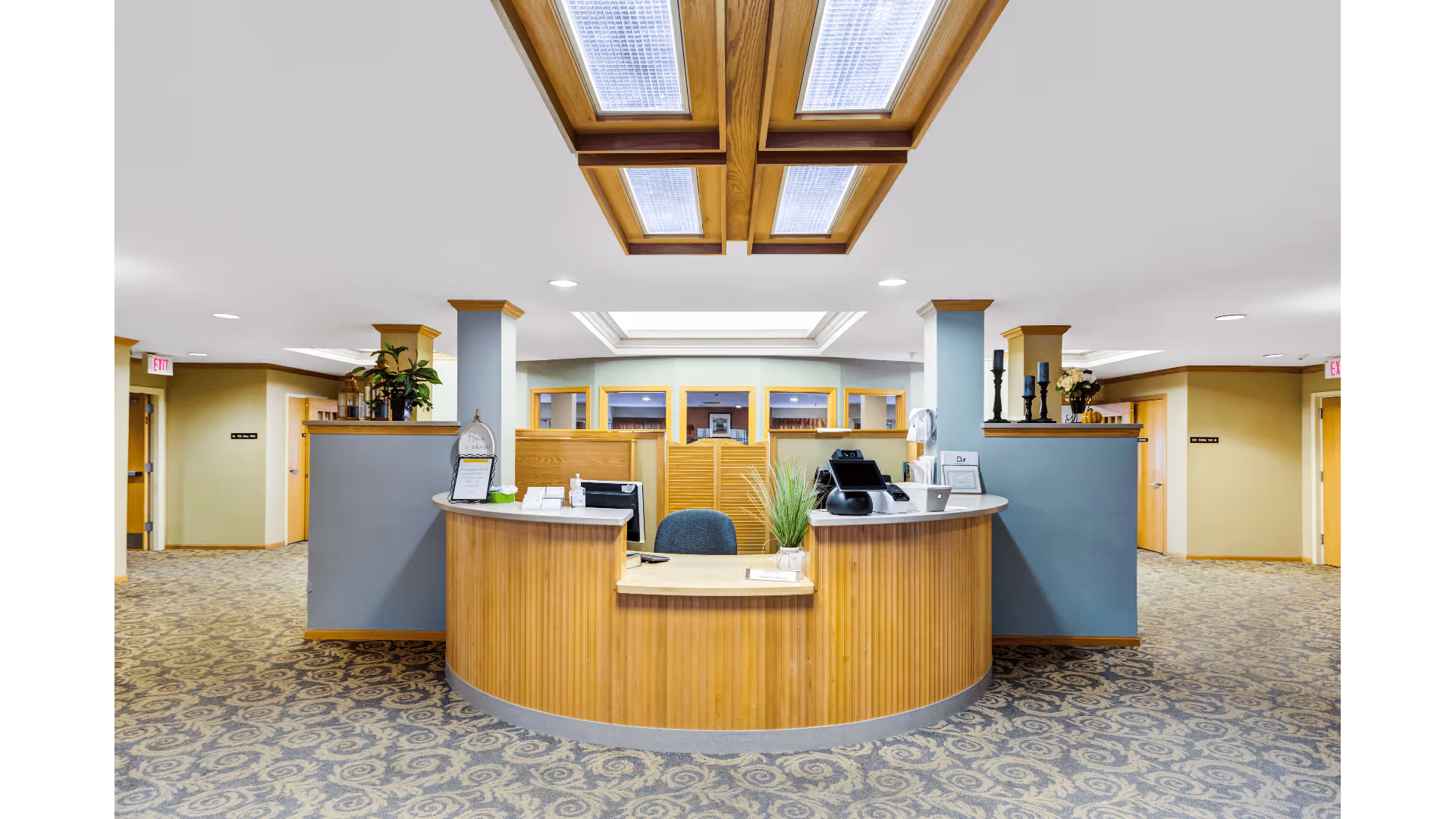 Reception desk area inside Dimensions Living Appleton facility with a curved wooden counter, office chair, computer monitors, decorative plants, and soft overhead lighting. The surrounding area has patterned carpet flooring, light-colored walls, and multiple doorways leading to other rooms.
