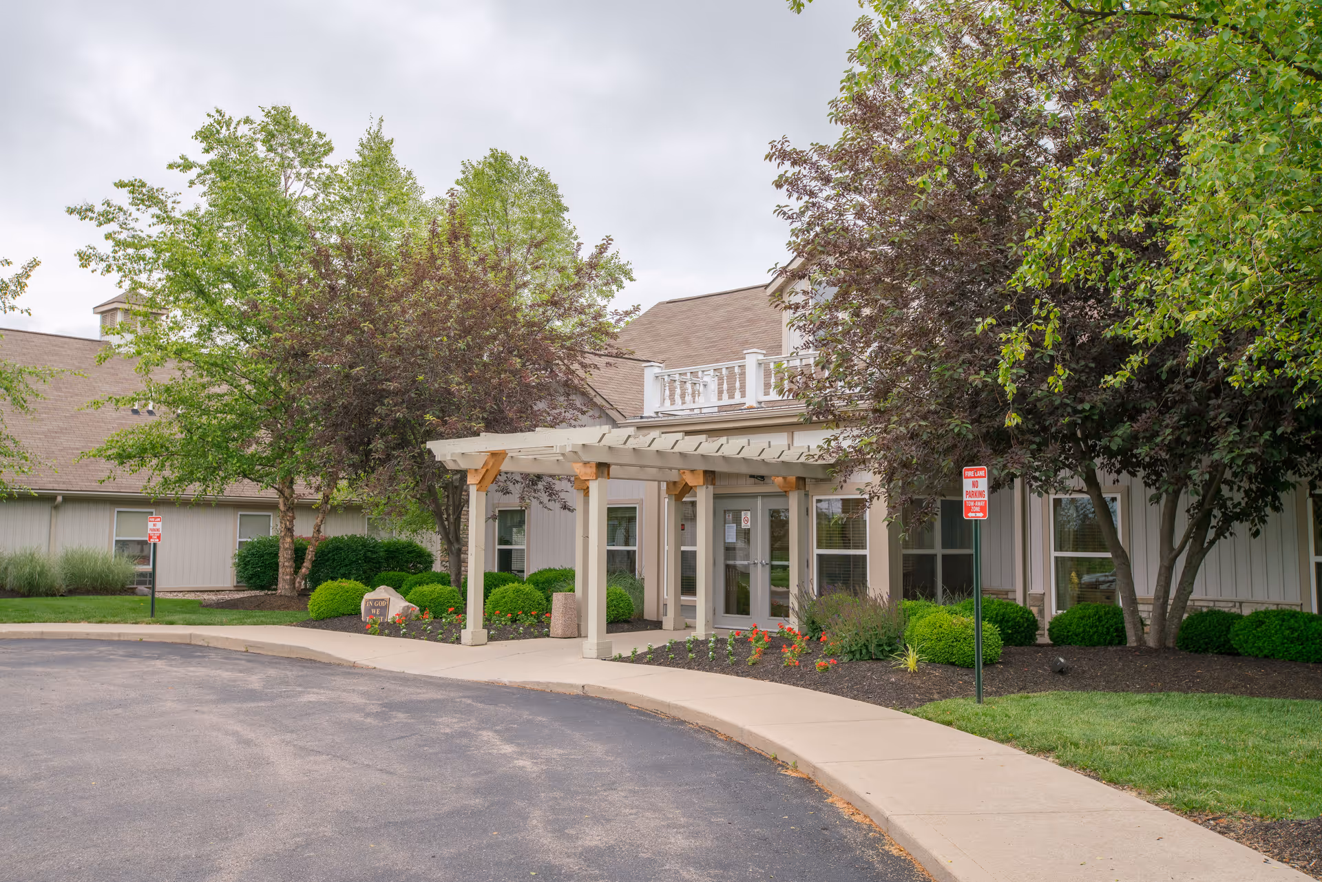 Exterior view of the entrance to Golden Years Nursing Center with a covered walkway, surrounded by green trees, shrubs, and a well-maintained garden bed. The building has a beige facade and a paved driveway leading to the entrance.