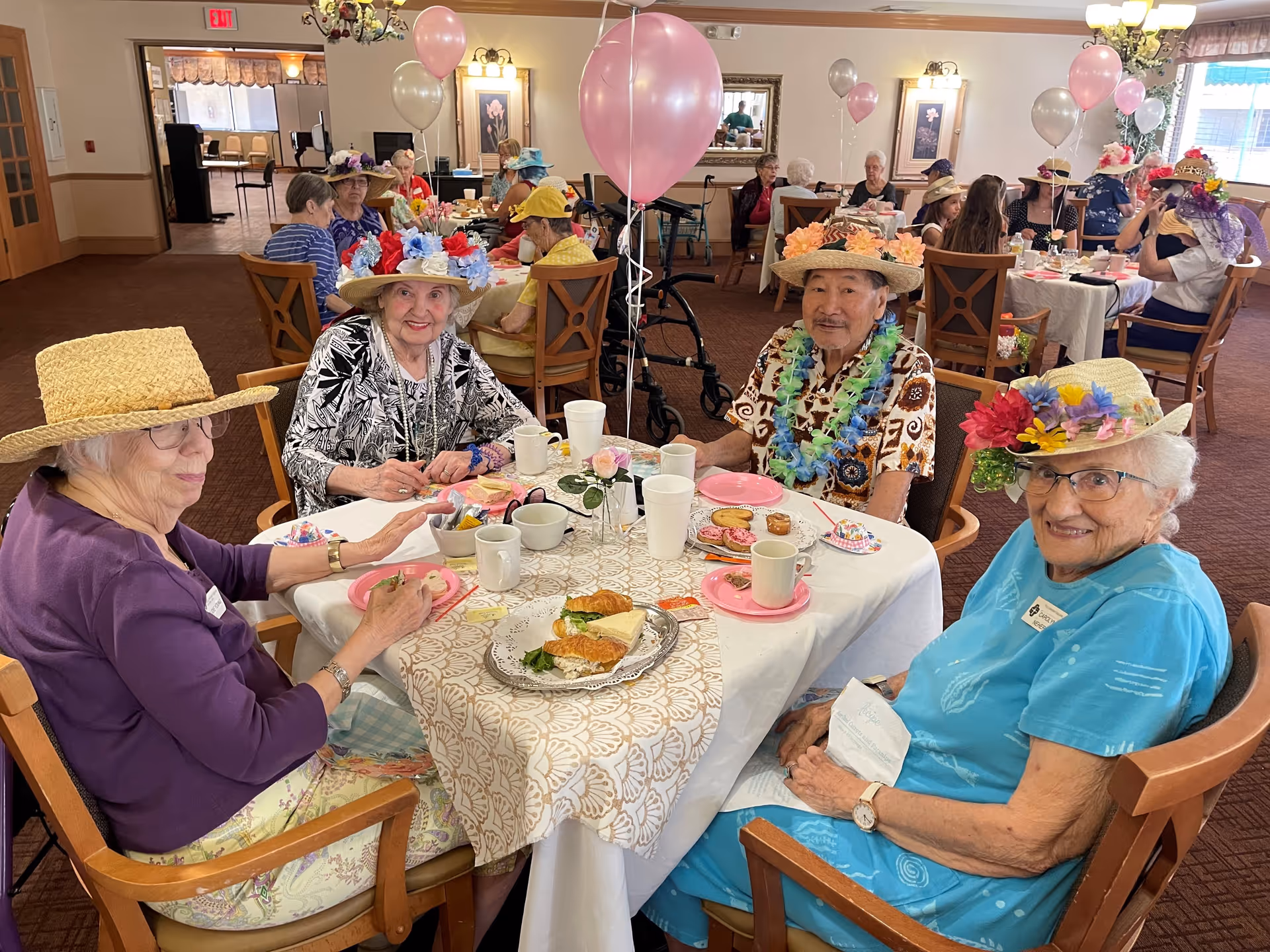 A group of seniors wearing decorative hats sit around a table with plates, cups and balloons in a festive dining room.