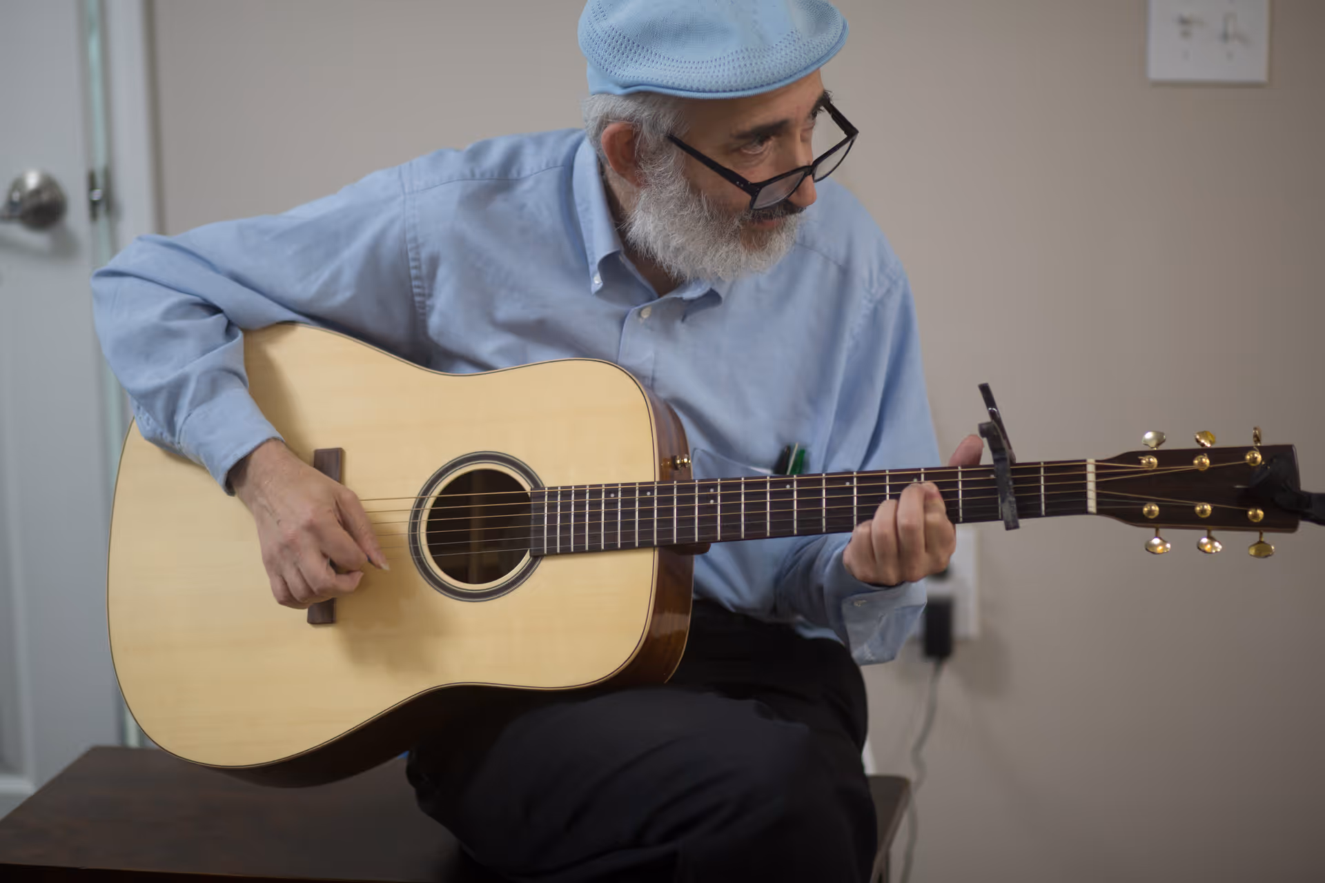 An elderly man wearing a light blue shirt and matching beret is sitting indoors playing an acoustic guitar. He is looking to his left and wearing glasses.