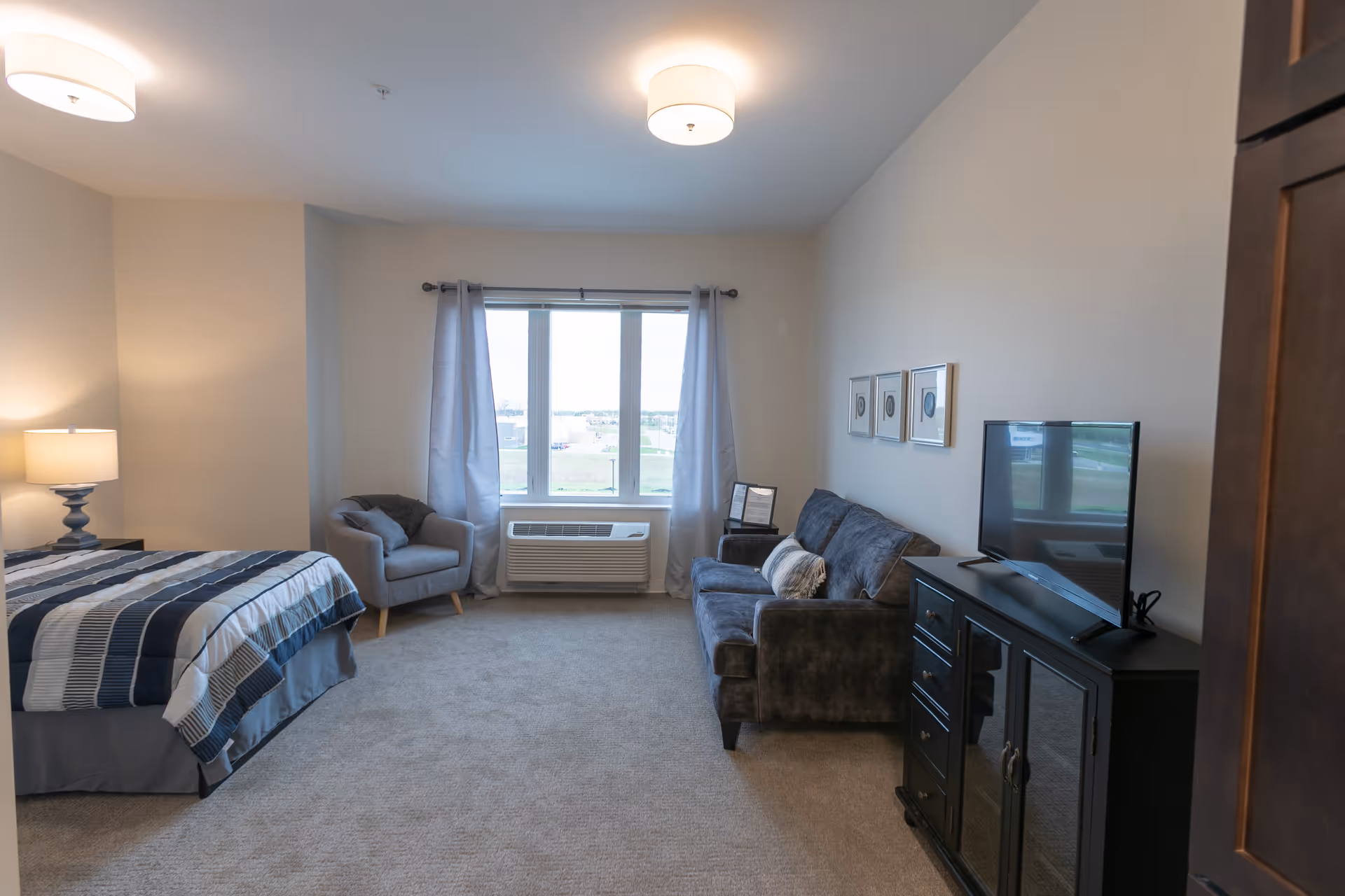A spacious bedroom with a bed covered in a blue and white striped comforter, a bedside table with a lamp, a gray armchair near a window with light gray curtains, a dark gray sofa with a pillow, a TV on a black cabinet, and three framed pictures on the wall.