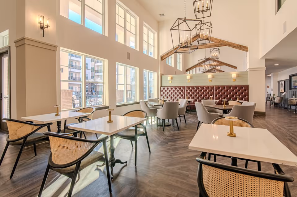 Bright and spacious dining area with multiple tables and chairs, large windows letting in natural light, high ceilings with exposed wooden beams, and modern chandeliers. The room features a red tufted bench along one wall and a wood floor.
