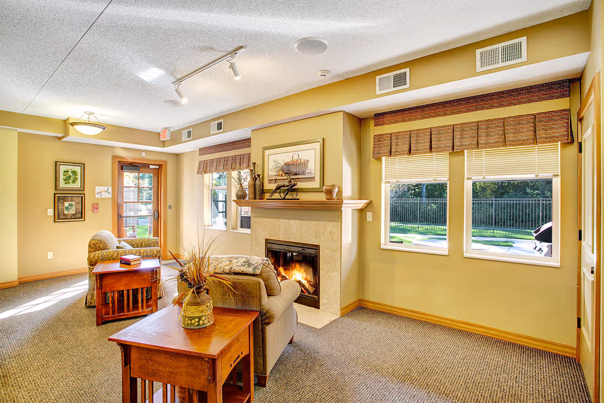 A cozy living room area in a senior living facility with beige walls and carpeted floor. The room features a lit fireplace with a mantel decorated with vases and a framed picture above it. There are two armchairs and wooden side tables, one with a decorative vase and the other with a book. Two windows with blinds and valances let in natural light, and a glass door leads outside to a patio area with greenery visible through the windows.