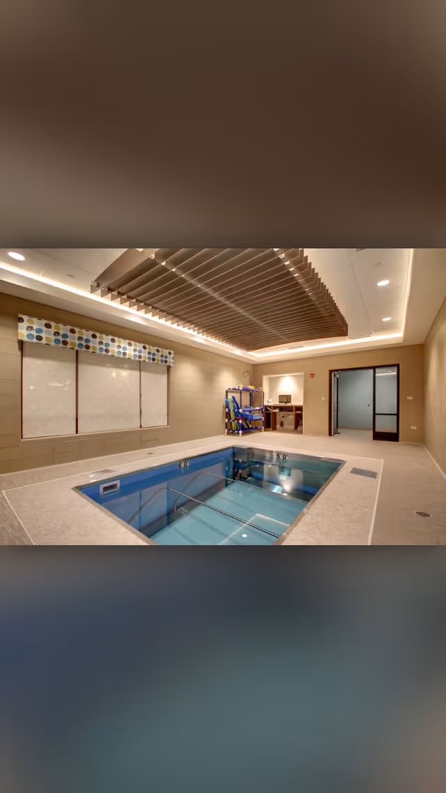 Well-lit indoor therapy pool in a room with decorative ceiling slats, pool lift equipment, and a glass doorway.