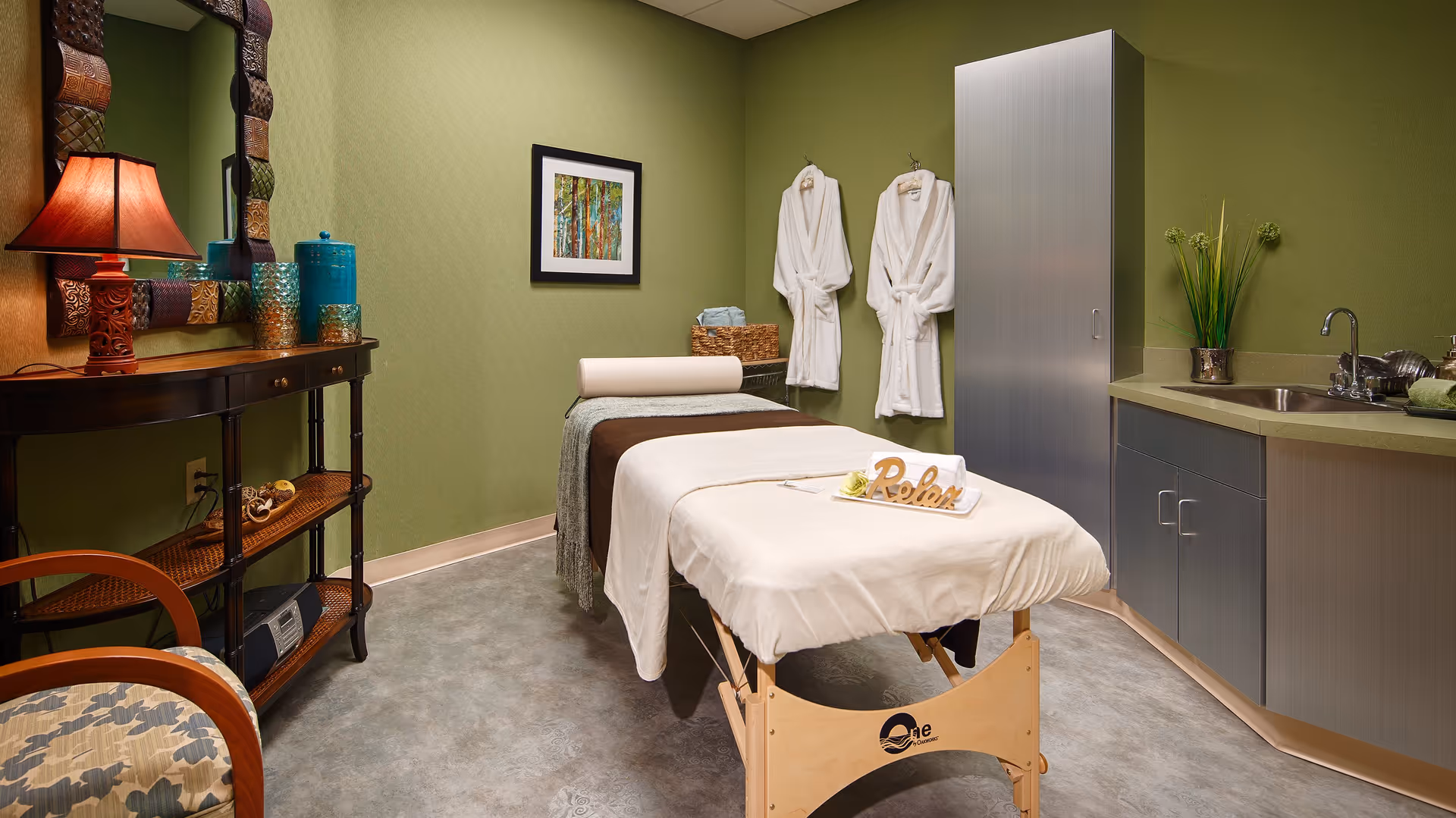 A serene massage therapy room with a massage table covered in white linens and a brown blanket. A wooden sign spelling 'Relax' is placed on the table. The room has green walls, a framed nature painting, two white robes hanging on the wall, a cabinet with a sink, and a decorative console table with a lamp and vases. A cushioned chair is partially visible in the foreground.