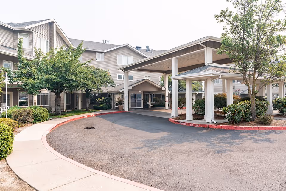 Exterior view of Maple Ridge Senior Living facility showing a covered entrance with white pillars, a circular driveway, and a multi-story building with beige siding and several windows surrounded by trees and shrubs.