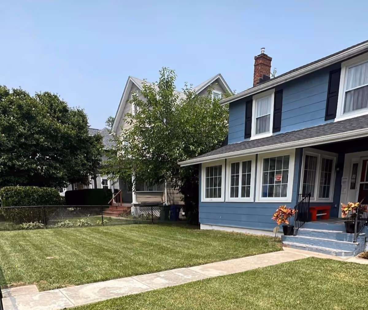 A blue two-story house with white trim and black shutters, featuring a front porch with steps leading up to it. The house has a well-maintained lawn with a concrete walkway and some potted plants near the porch. There are neighboring houses and trees in the background under a clear sky.