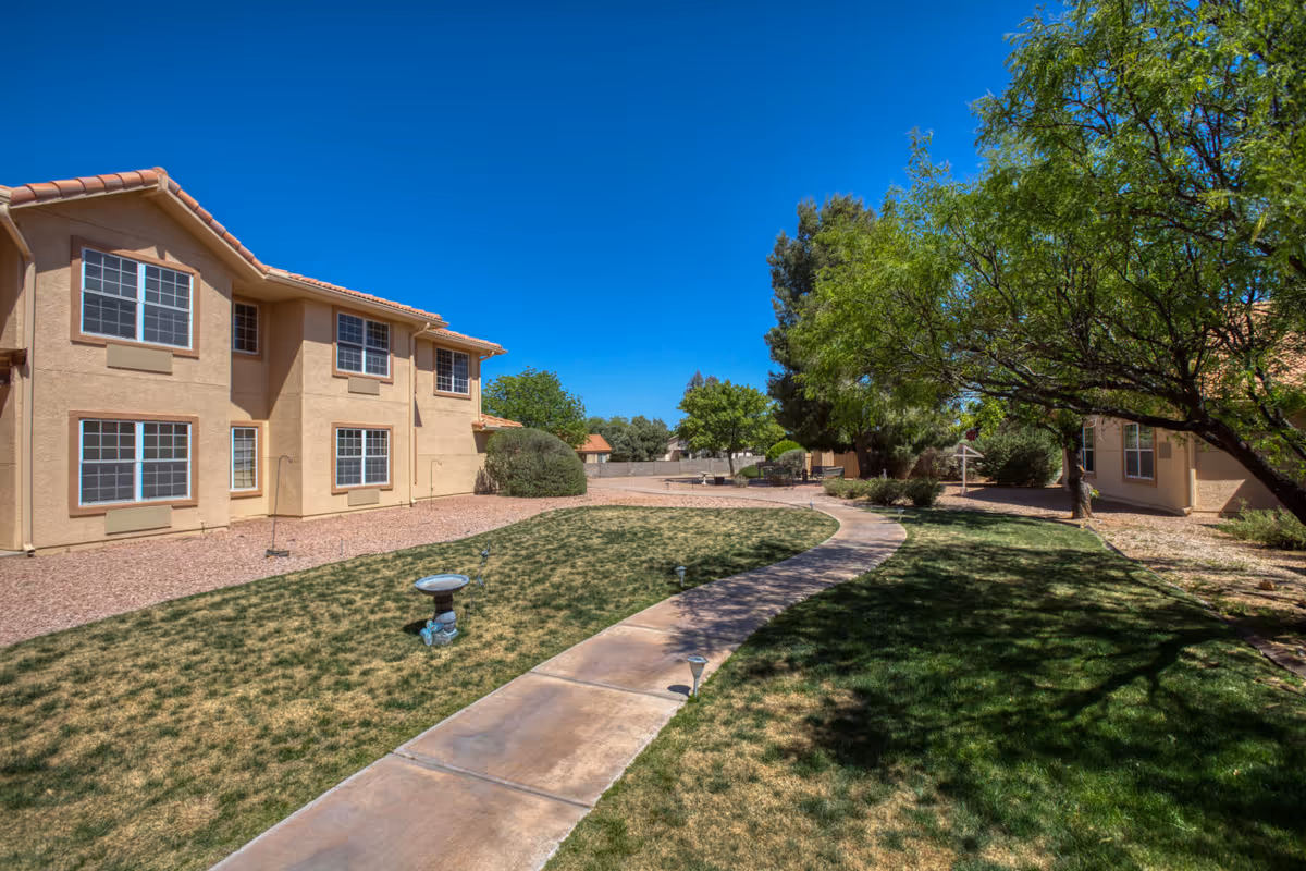 Sunlit courtyard with a curved walkway, grassy lawn, trees, and a two-story beige building.