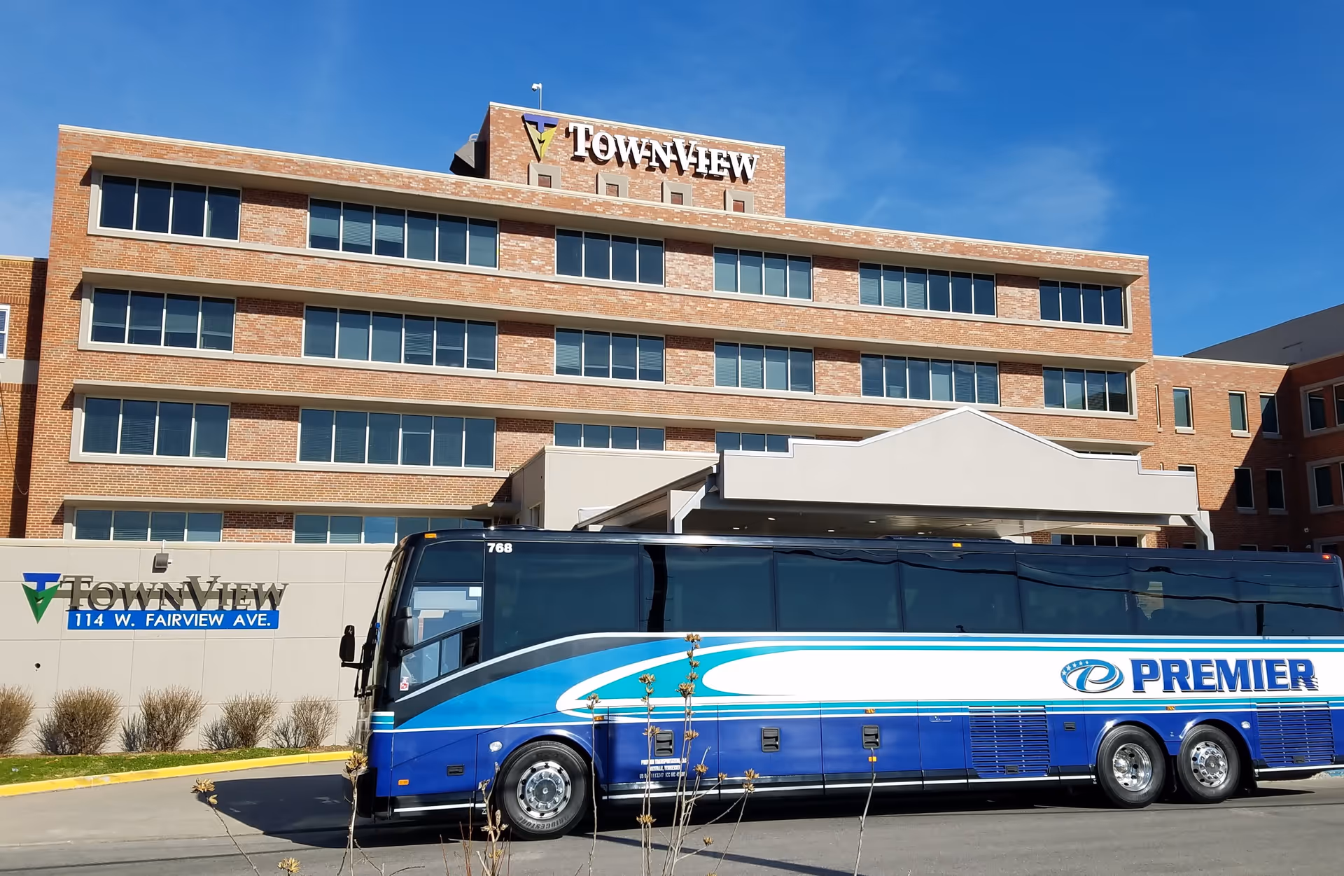 Exterior view of the TownView Senior Living building, a multi-story brick structure with many windows. A large blue and white Premier bus is parked in front of the building under a covered entrance. The sky is clear and blue.