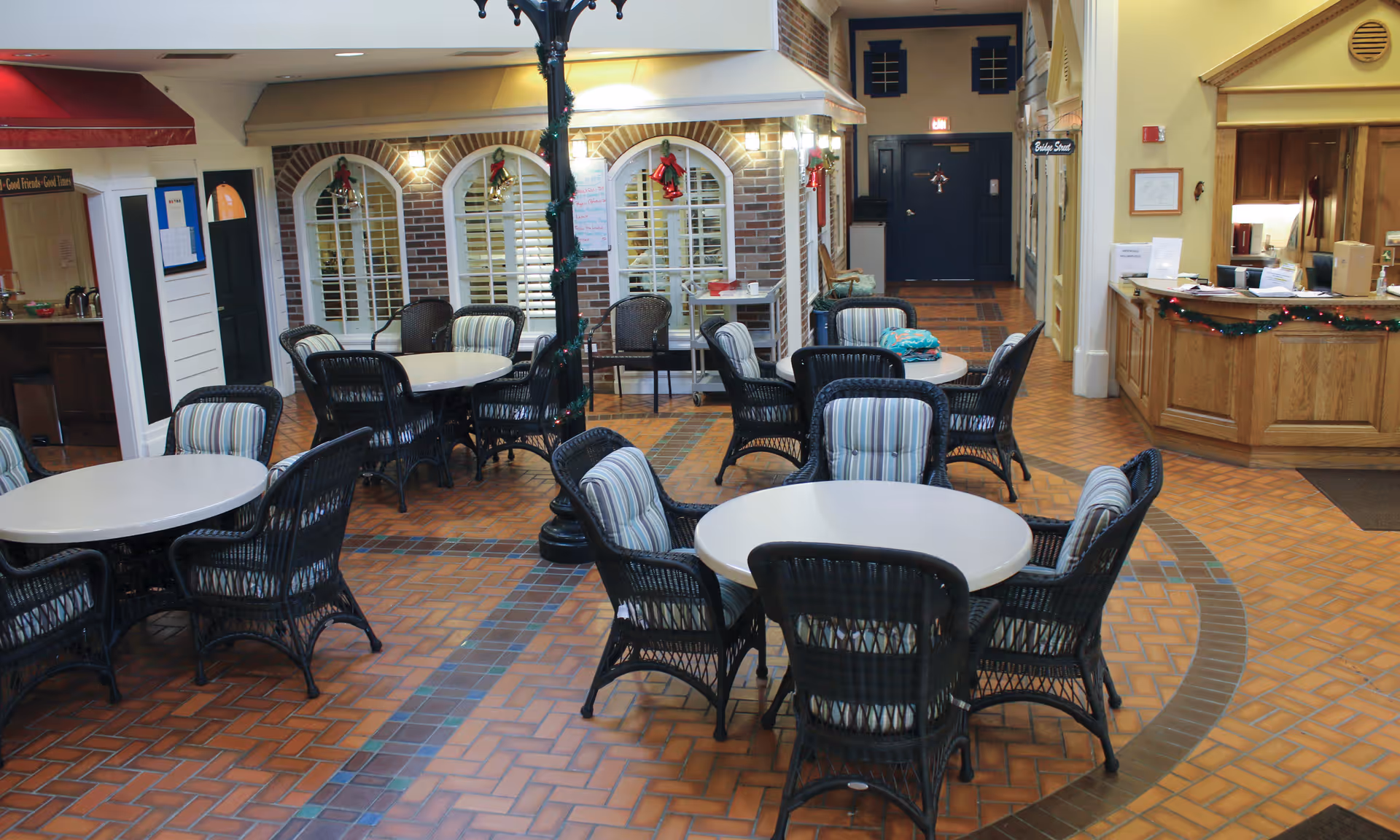Indoor common area with several round tables surrounded by black wicker chairs with striped cushions. The floor is tiled with a brick pattern, and there are decorative elements such as a lamppost wrapped with garland and holiday decorations on the walls and windows. A reception desk is visible on the right side of the image.