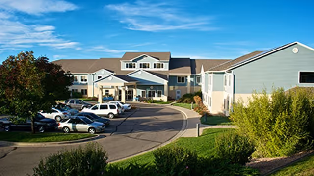 Exterior view of a senior living facility with a curved driveway, parked cars, landscaped greenery, and a blue sky.