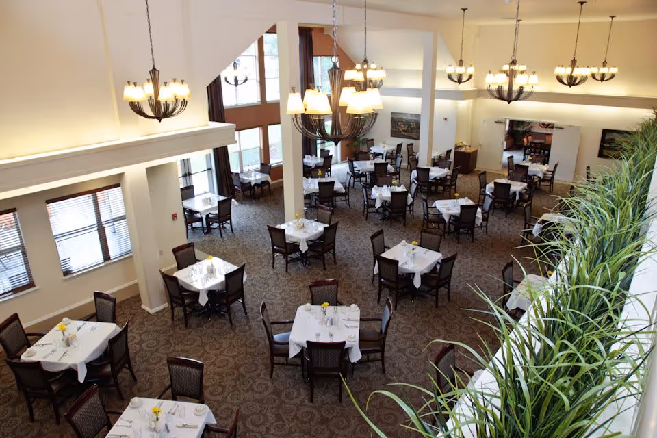 A spacious dining room with multiple tables covered in white tablecloths, each set with plates, cups, and yellow flowers in small vases. The room features large windows with blinds, several chandeliers hanging from the ceiling, and a patterned carpet. There are green plants along a ledge on the right side of the image.