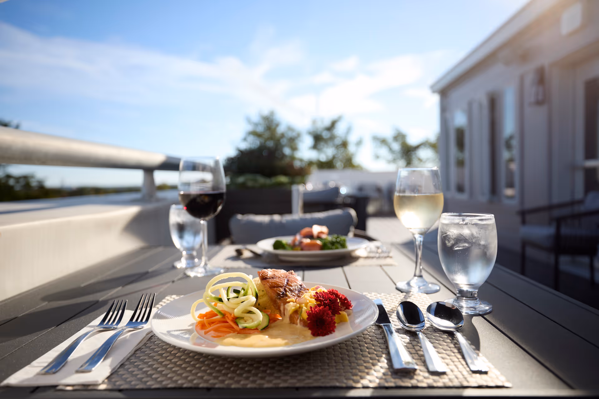 Outdoor dining table set with plates of food, glasses of red and white wine, and water glasses on a sunny patio with a railing and greenery in the background.