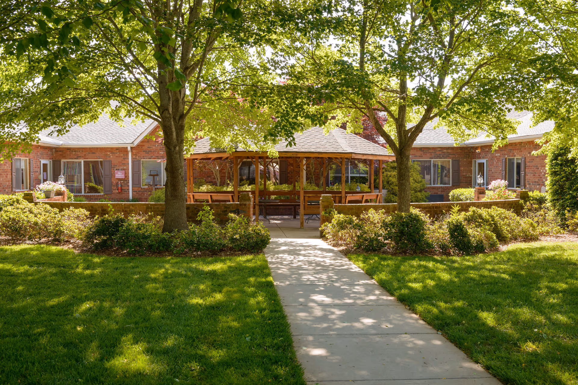 A sunny outdoor garden area with a concrete pathway leading to a wooden gazebo surrounded by green trees, bushes, and well-maintained grass. The background shows a brick building with windows and flower pots.