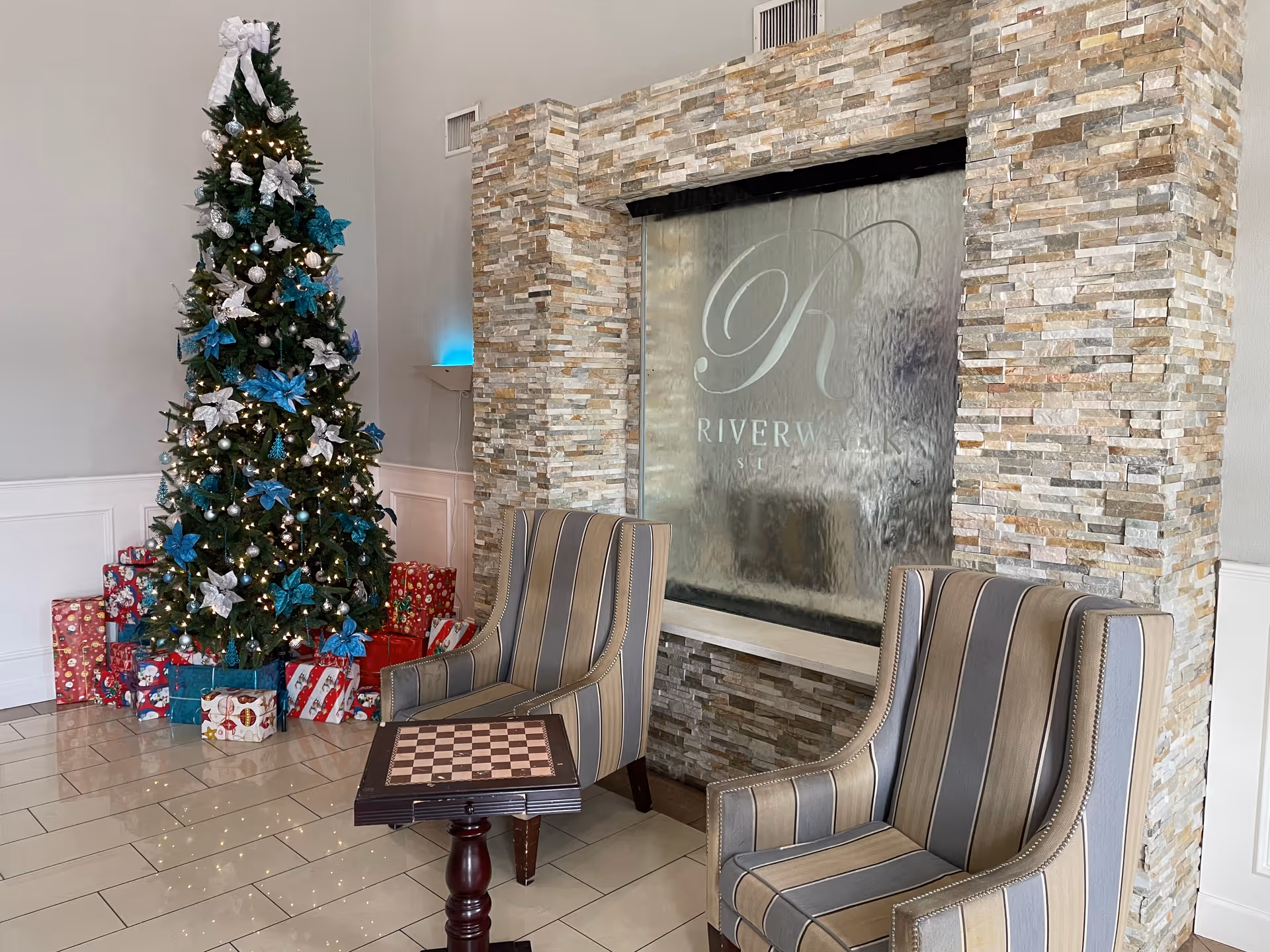 Lobby seating area with two striped chairs, a small chess table, a decorated Christmas tree with presents, and a stone water feature displaying the Riverwalk name.
