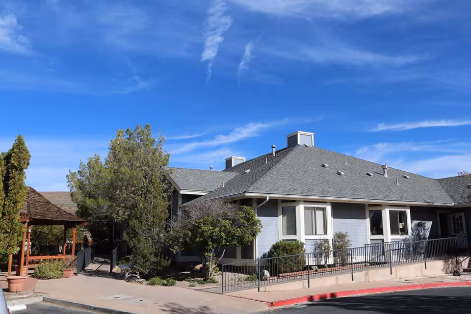 Exterior view of a single-story assisted living facility building with gray siding and a gray shingled roof under a blue sky with some clouds. There are trees and shrubs around the building, a paved walkway, and a wooden gazebo on the left side.