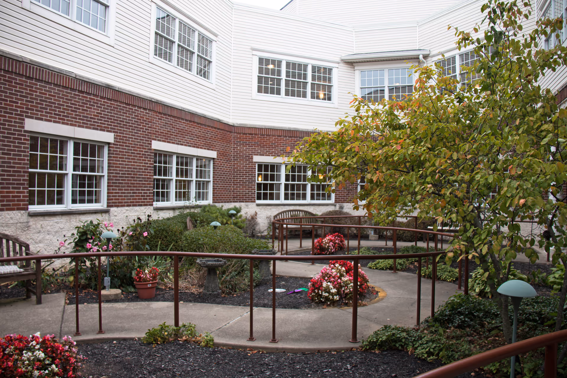 Outdoor courtyard area with a paved walking path, metal handrails, benches, flower beds with blooming flowers, shrubs, and a tree. The courtyard is surrounded by a multi-story building with brick and white siding exterior and multiple windows.